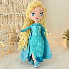 a crocheted doll with blonde hair and blue dress standing next to a plant