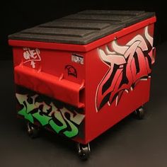 a red trash can with graffiti on it's lid and wheels, sitting in front of a black background