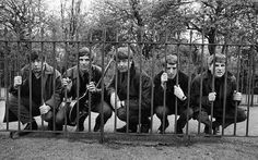 black and white photograph of five men behind bars in front of trees with one holding a guitar