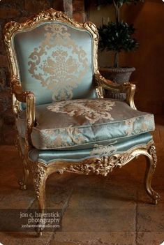 an ornate gold and blue chair sitting in front of a potted plant on a stone floor