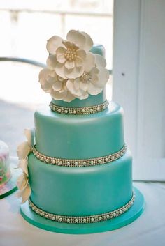 a three tiered blue cake with white flowers on it's side and pearls around the edges