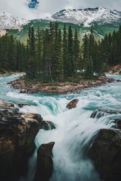 a river running through a forest filled with rocks and trees next to snow covered mountains