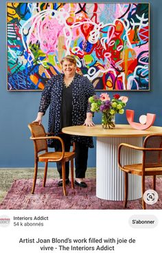 a woman standing in front of a table with flowers on it next to a large painting