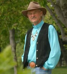 a man wearing a cowboy hat and blue shirt standing in front of trees with his hands on his hips