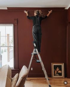 a woman is standing on a ladder and painting the wall in her living room with red walls