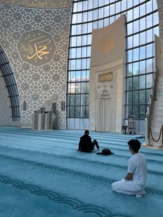 two men sitting on the ground in front of a building with large windows and arabic writing