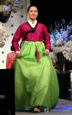 a woman is walking down the runway wearing a green skirt and pink shirt with an elaborate flower arrangement behind her