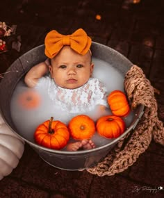 a baby sitting in a tub with pumpkins on the ground next to it and wearing an orange bow