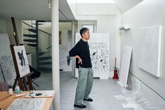 a man standing in an art studio looking up at the skylight above him and drawing on canvases behind him