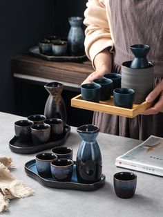 a woman is holding a tray with cups and vases on it while another person holds a book in the background