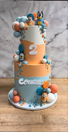 a three tiered cake decorated with cartoon characters and balloons is on a table in front of a marble wall