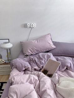 an unmade bed with purple sheets and pillows on it in a white walled room