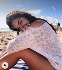 a woman sitting on top of a sandy beach next to the ocean with her hand under her face