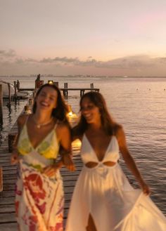 two beautiful women standing next to each other on a pier near the water at sunset