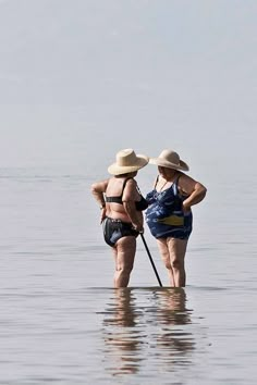 two women standing in the water with their backs to each other, wearing hats and bathing suits