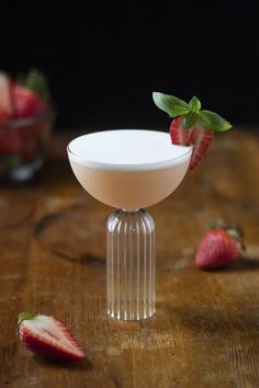 a drink in a coupe glass with strawberries on the table next to it and another bowl full of strawberries