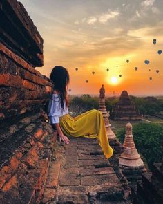 a woman sitting on top of a stone wall near hot air balloons in the sky