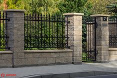 an iron fence with stone pillars and columns on the side of a street in front of trees