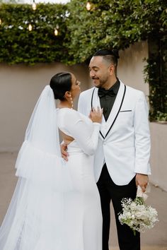 a bride and groom standing next to each other in front of a wall with greenery