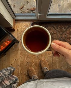 a person holding a cup of tea in front of an open fire place with their feet on the floor