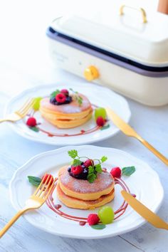 two white plates topped with pancakes covered in berries and raspberries next to a fork