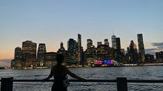 a woman standing on the edge of a pier looking at the city lights and skyscrapers