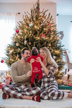 a man and woman are sitting in front of a christmas tree holding a small child