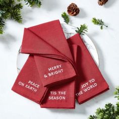 four red napkins with merry and bright on them sitting next to pine cones, evergreen branches