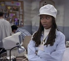 a woman with dreadlocks and a hat standing in front of a computer desk