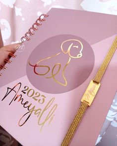 a person holding a pink notebook with gold foil on it and a dog sticker attached to the cover