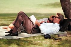 two men laying on a blanket under a tree talking on their cell phones and reading books