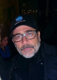 a man with glasses and a hat standing next to another man in a black jacket