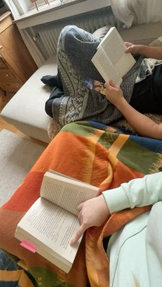 two people sitting on a bed with books in their hands and one person holding an open book