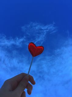 a hand holding a heart shaped lollipop in front of a blue sky with clouds