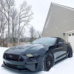 a black mustang parked in the snow next to a garage with trees and a house behind it