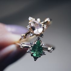 someone is holding an engagement ring with two pear shaped diamonds and green emeralds on it