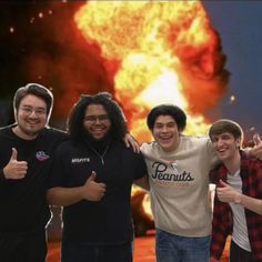 four young men are posing in front of a large explosion with their thumbs up and one is giving the thumbs up sign