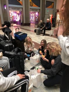 four women sitting on the floor eating and drinking in an airport terminal with their luggage