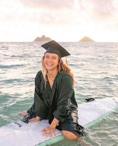 a woman sitting on top of a surfboard in the ocean wearing a graduation cap