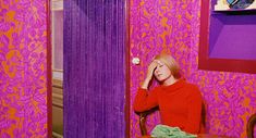 a woman sitting at a table in front of a pink wallpapered room with purple curtains