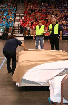 two men in yellow vests are setting up beds on a stage with people watching