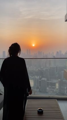 a woman standing in front of a window looking out at the cityscape and sunset