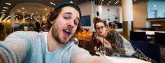 a man and woman taking a selfie in an office setting with people sitting at tables