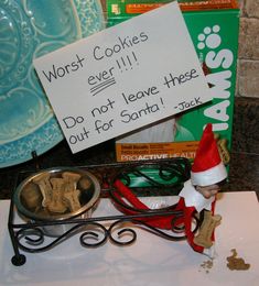 the elf is sitting in front of some cookies on top of a table with a sign that says, worst cookies ever will do not leave these out for santa