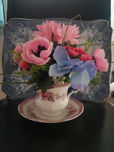 pink and blue flowers are in a teacup on a table with a decorative plate