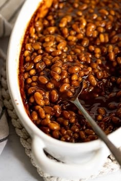 a white bowl filled with baked beans on top of a table next to a spoon