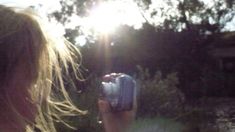 a woman taking a photo with her cell phone in the sun shining through trees and bushes