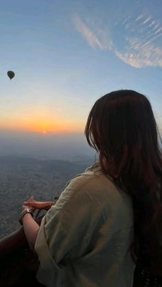 a woman sitting on top of a hill looking at a hot air balloon in the sky