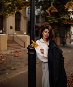 a woman standing next to a black pole on the side of a road with an autumn leaf in her hand