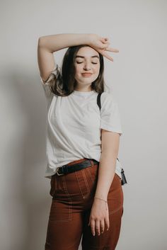 a woman in brown pants and a white t - shirt is holding her hands behind her head